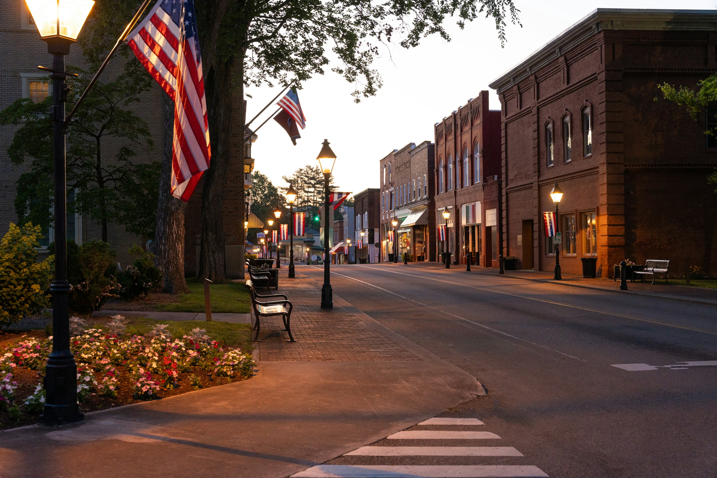 Downtown Redstone's Main Street at dusk, lined with brick storefronts, string-lit trees and American flags.